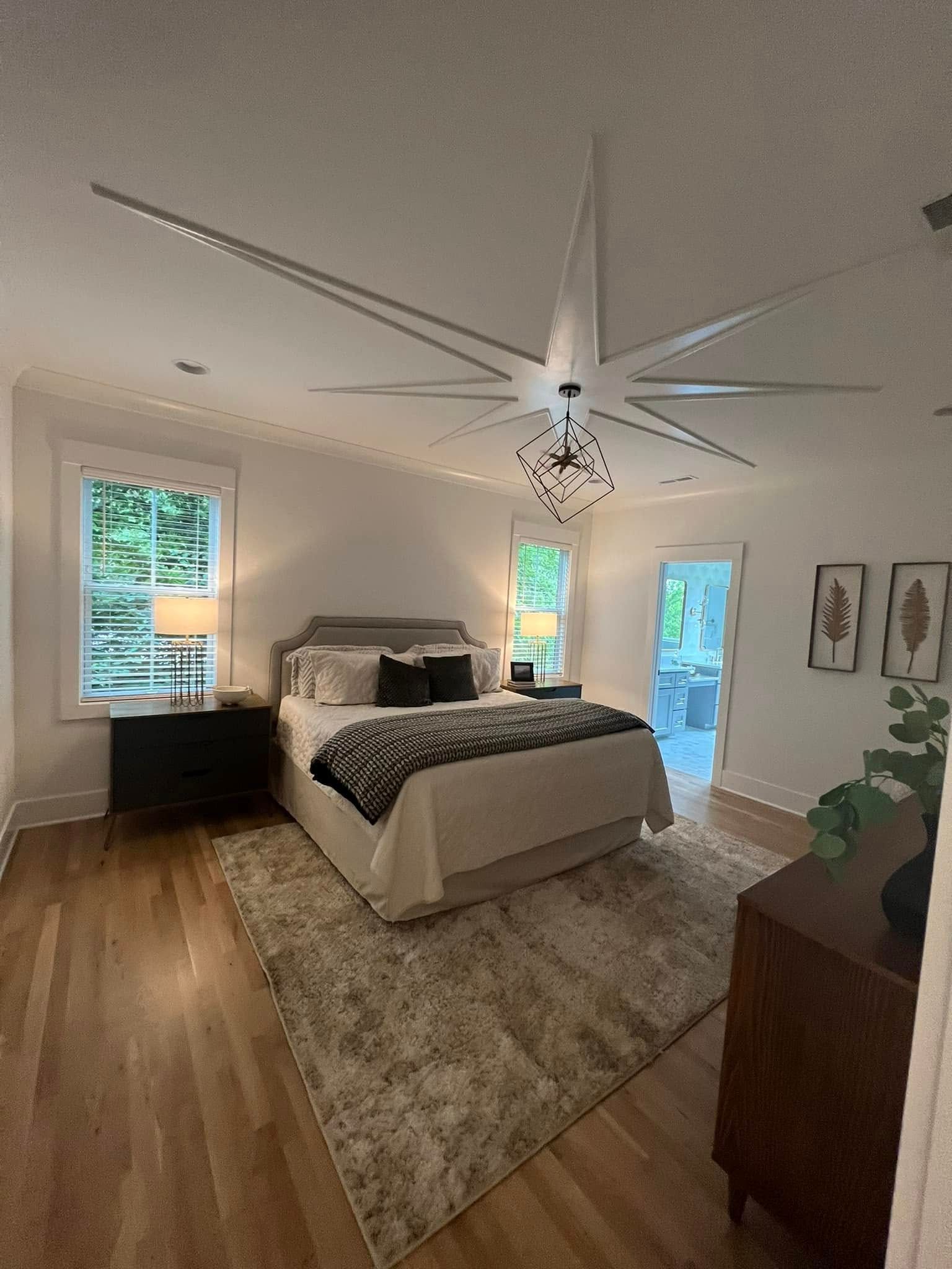 Bright modern bedroom with white walls, a bed, wood floor, large starburst ceiling light, and windows with greenery outside