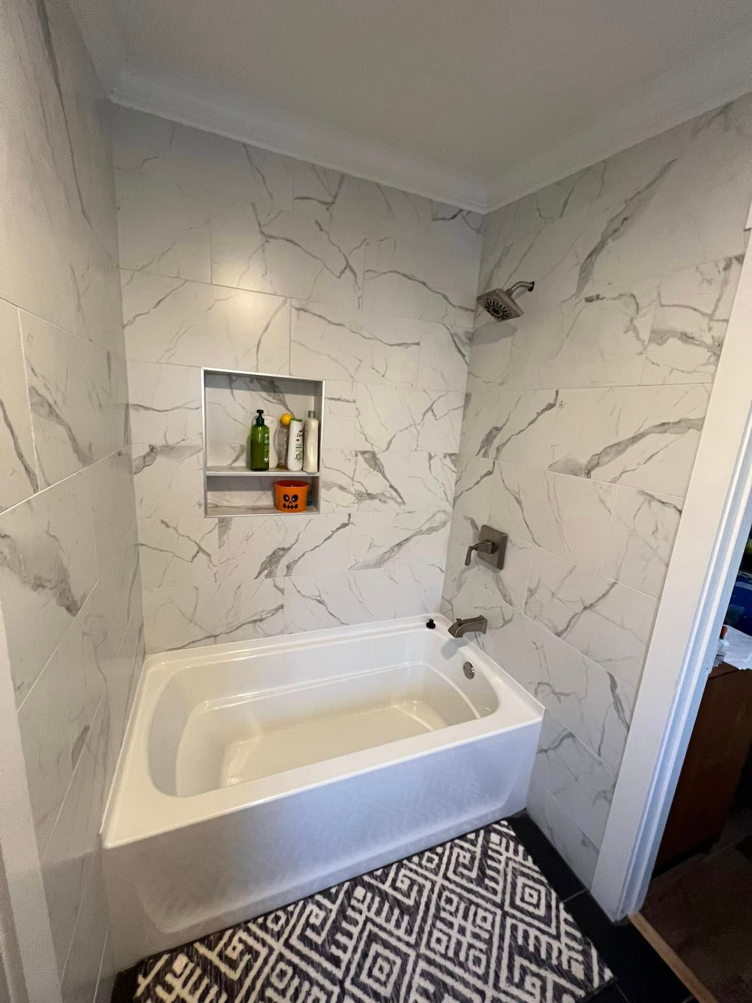 White marble-tiled bathroom with a built-in tub, patterned rug, and recessed shelf holding bottles and decor.