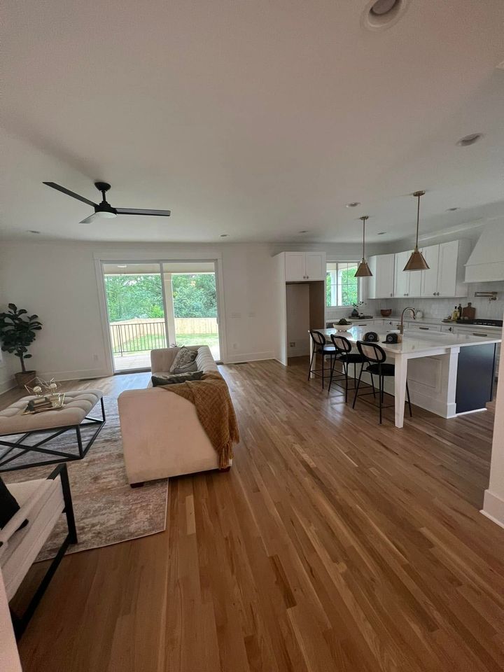 Bright open living room and kitchen with wood floors, white walls, ceiling fans, and large windows