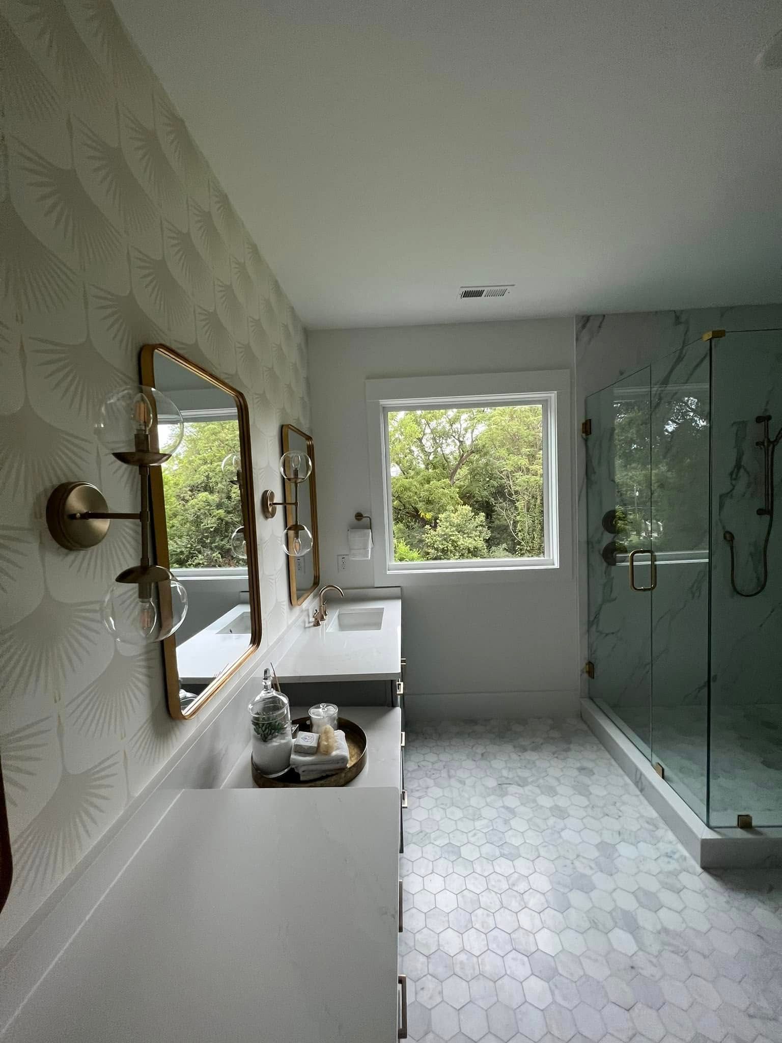 Modern bathroom with double sink vanity, gold mirrors, walk-in glass shower, and window.