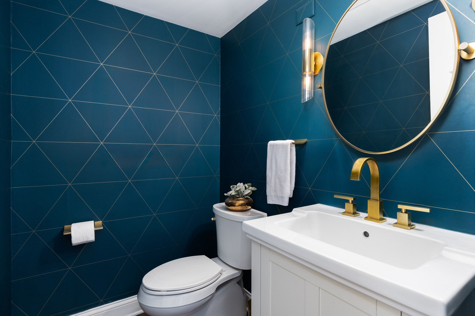 Small teal bathroom with gold fixtures, white vanity, toilet, round mirror, and wall sconce.
