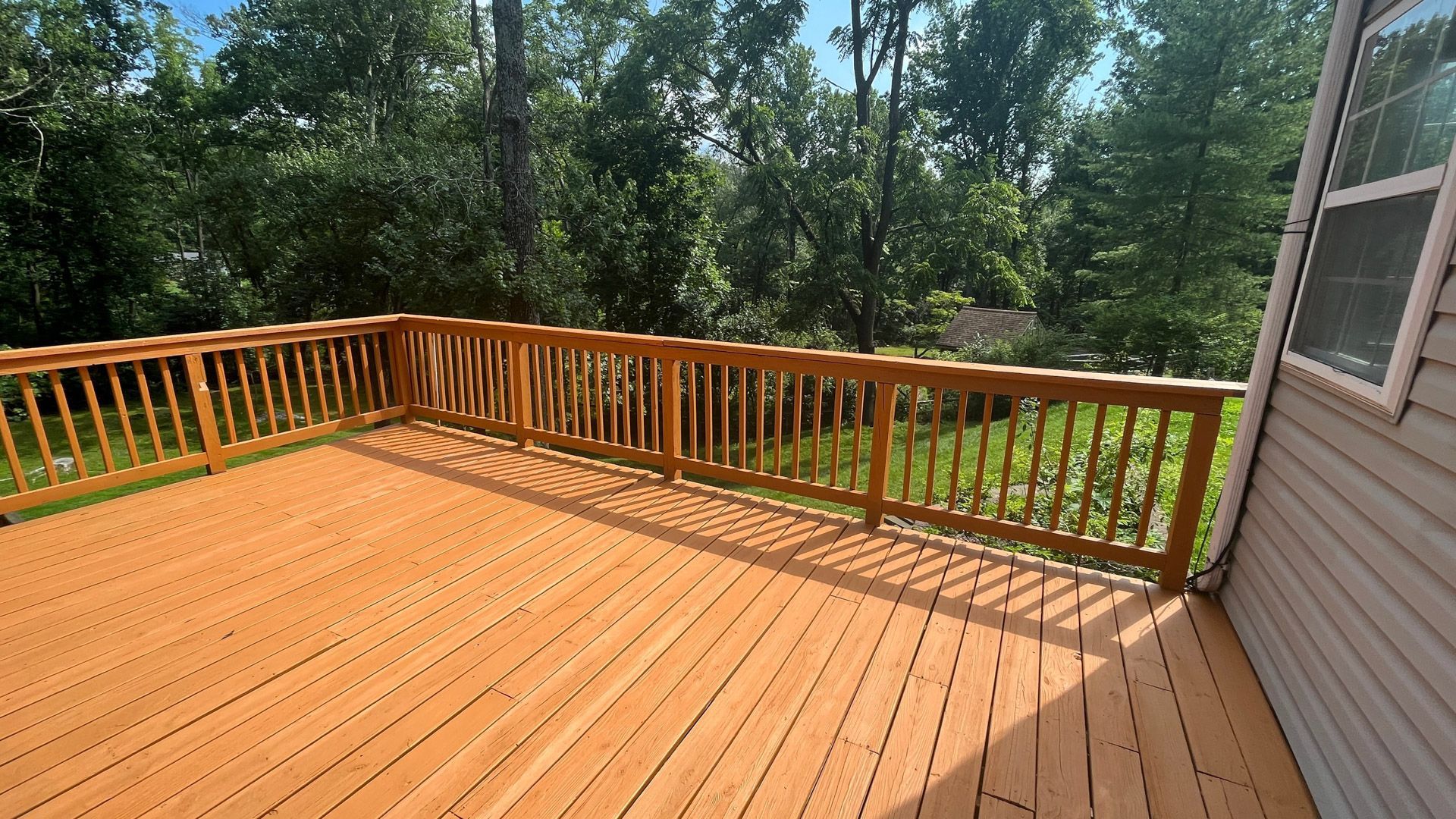 Sunny wooden deck with railing overlooking a grassy, tree-lined yard