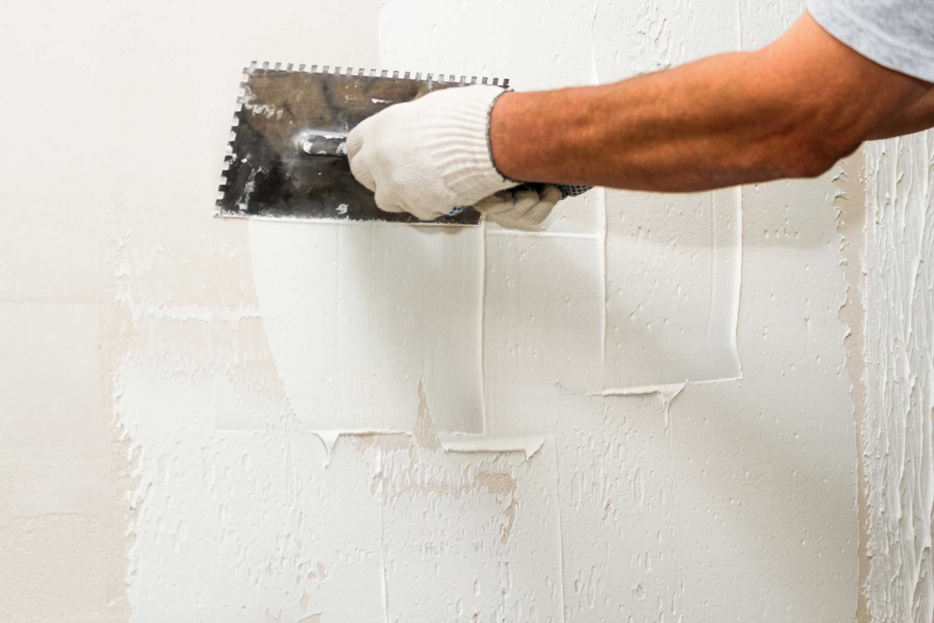 Gloved hand using a trowel to smooth plaster on a white wall.