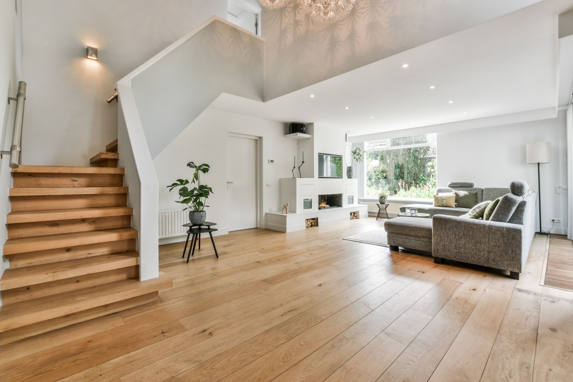 Bright open-plan living room with wooden floors, white staircase, and gray sofa by large windows
