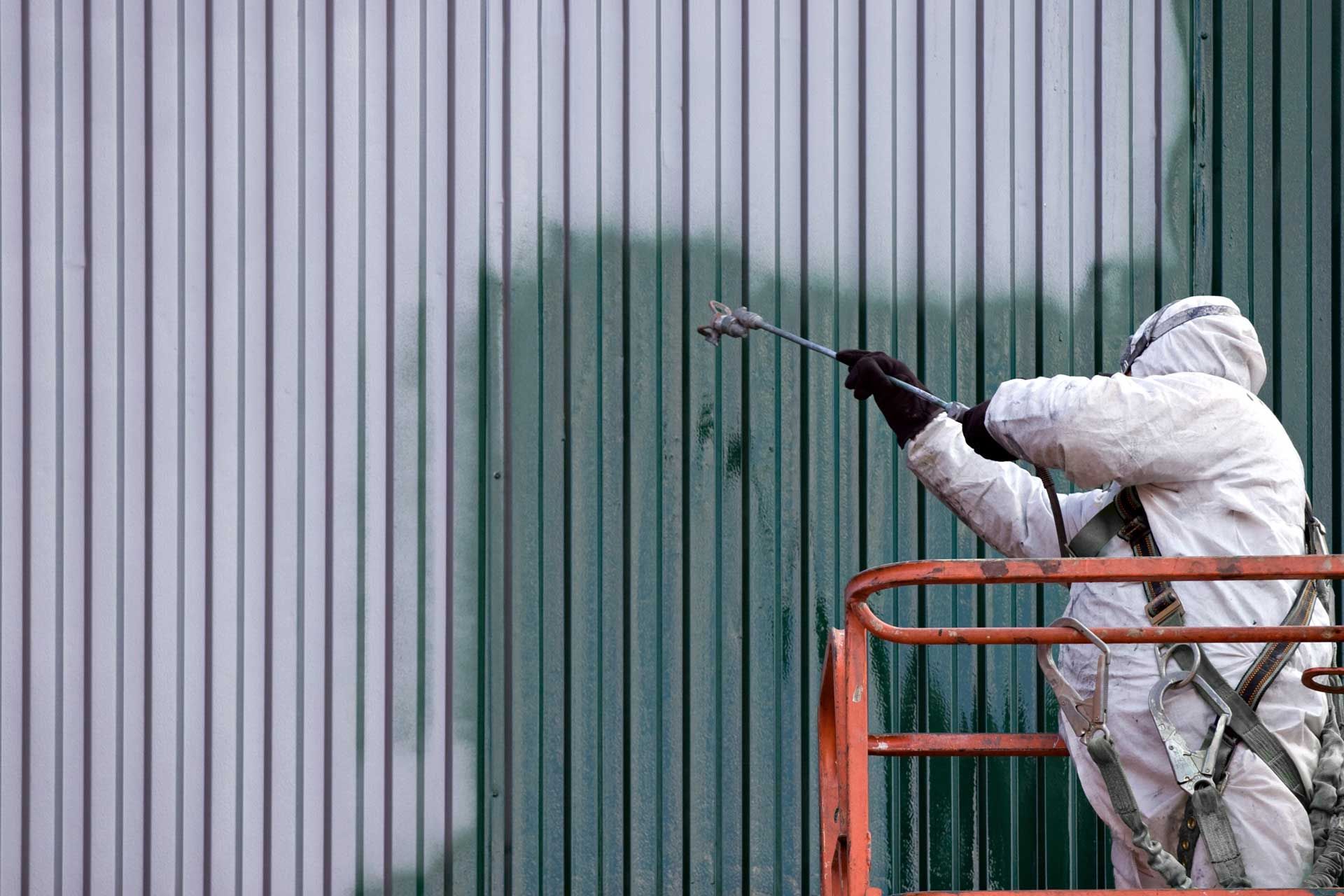 Worker in white protective suit spray-painting a corrugated wall from an orange lift platform
