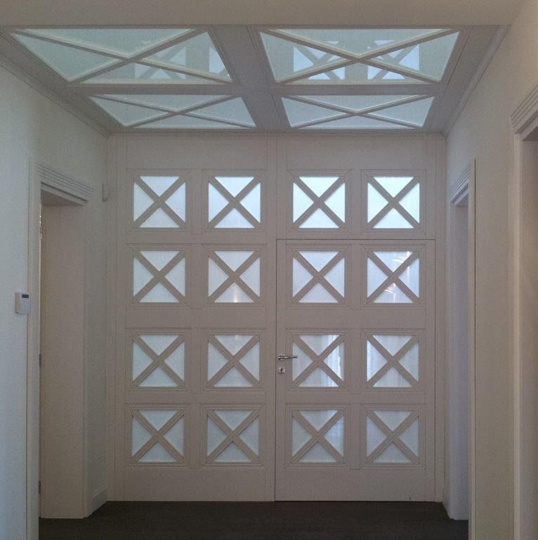 Corridoio bianco con doppia porta a pannelli con motivo a X e lucernari geometrici coordinati sul soffitto.