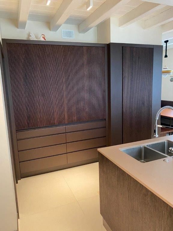 Cucina moderna con mobili in legno scuro, isola centrale con lavello e illuminazione a pavimento con piano di lavoro beige.