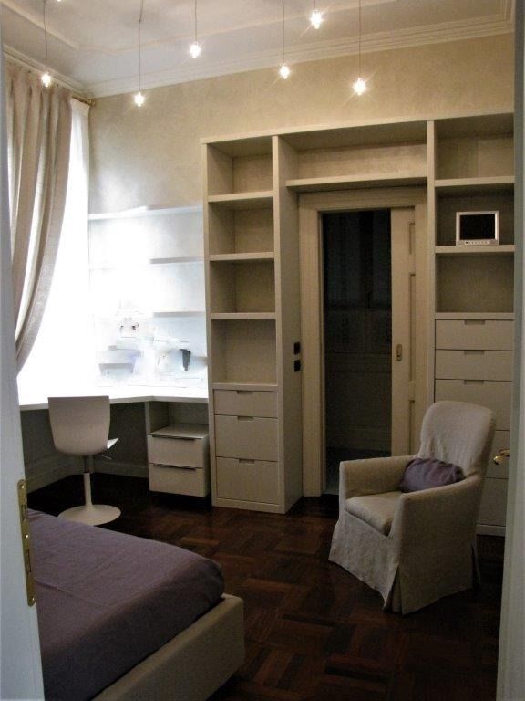 Piccola camera da letto beige con scrivania a incasso, mensole, cassetti, letto e sedia.