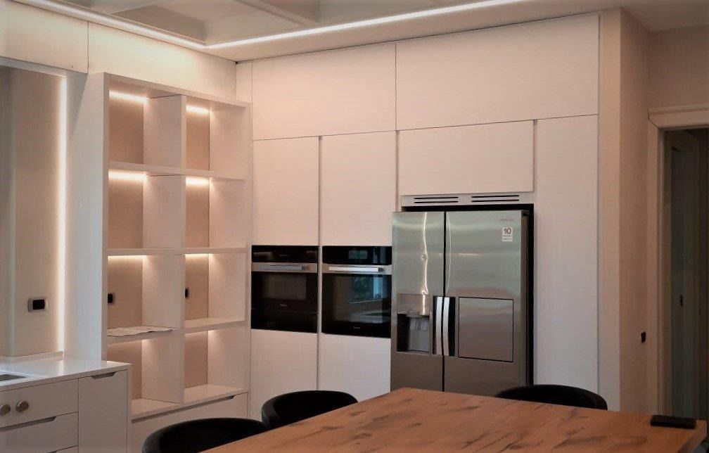 Cucina moderna con mobili bianchi, forni a incasso, frigorifero in acciaio inox e tavolo da pranzo in legno.