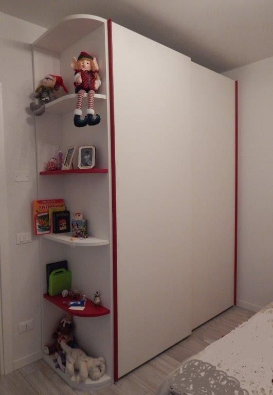 Armadio bianco a muro con ripiani rossi che espongono bambole e giocattoli nella cameretta di un bambino.
