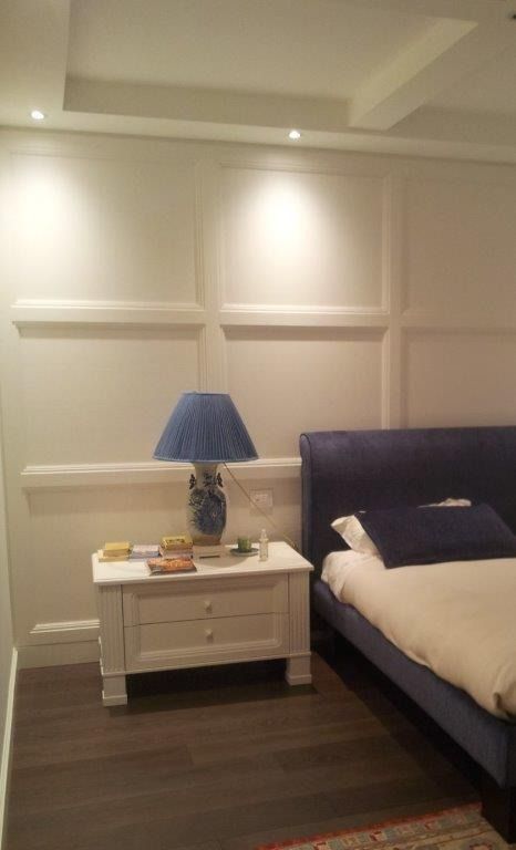 Elegante camera da letto con letto in legno scuro, comodino bianco, lampada blu e parete rivestita in legno color crema.