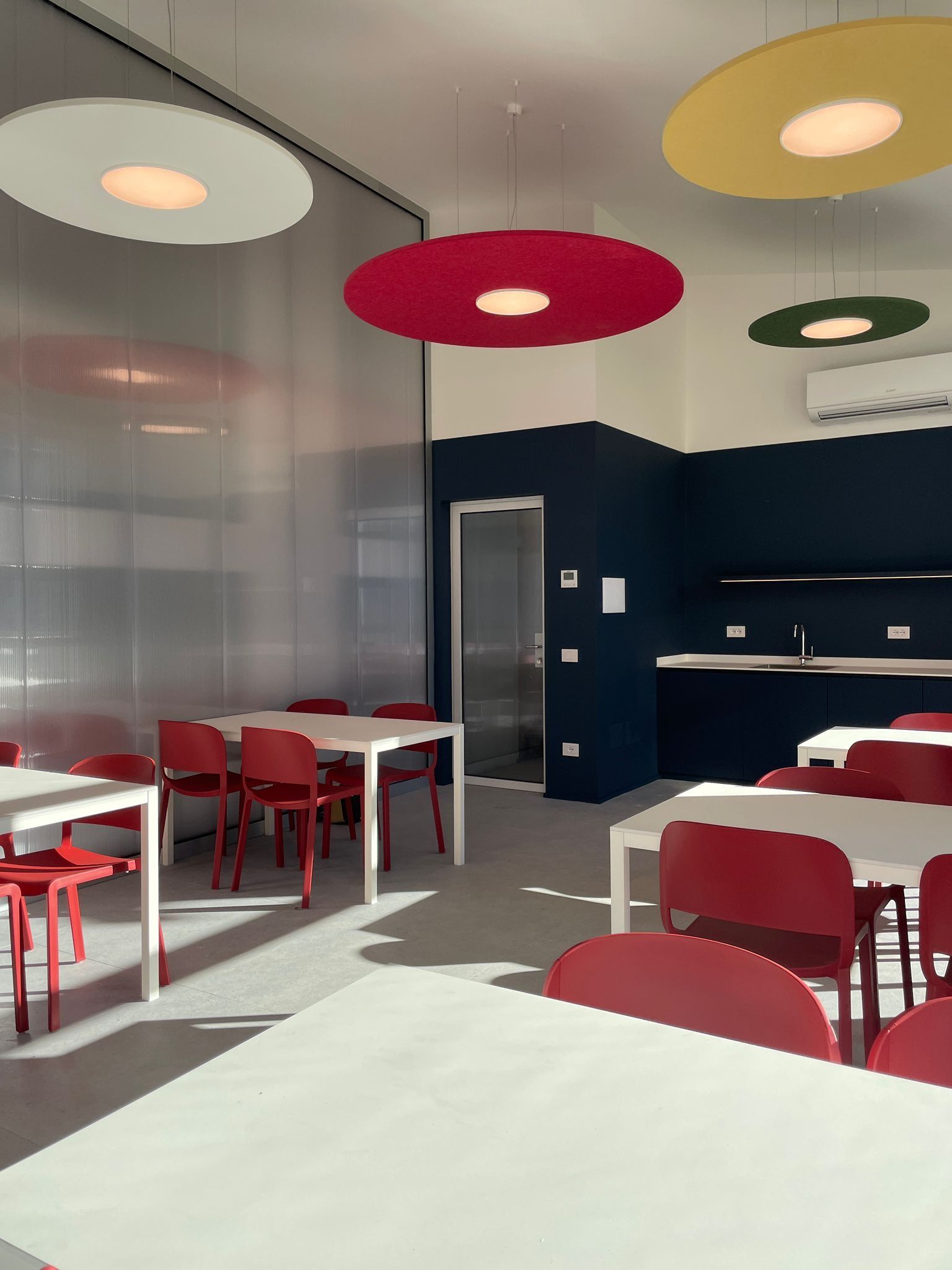 Interno moderno di un caffè con sedie rosse, tavoli bianchi e lampade a sospensione colorate.