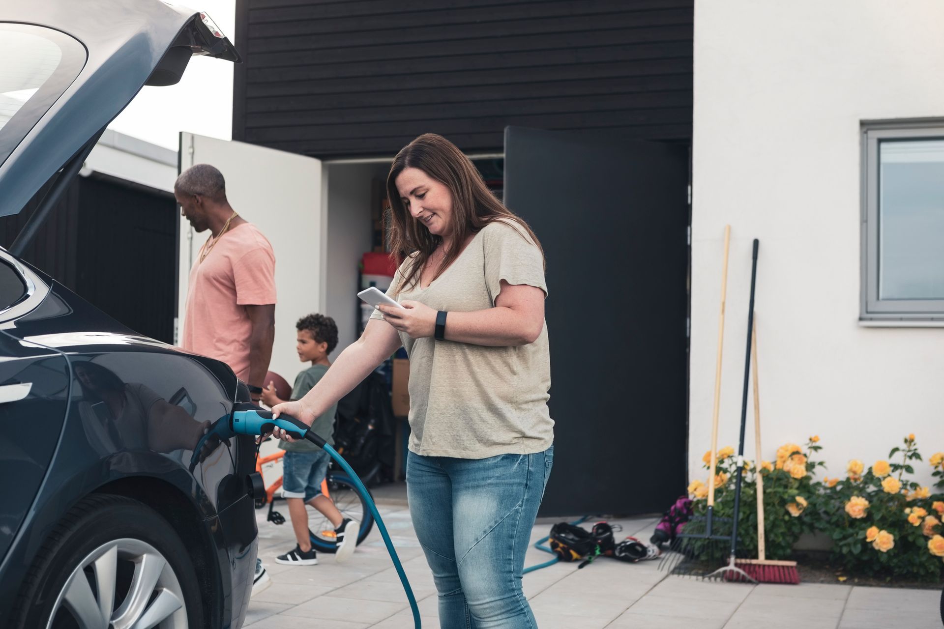 A mother using a smartphone while charging an electric car in the front yard