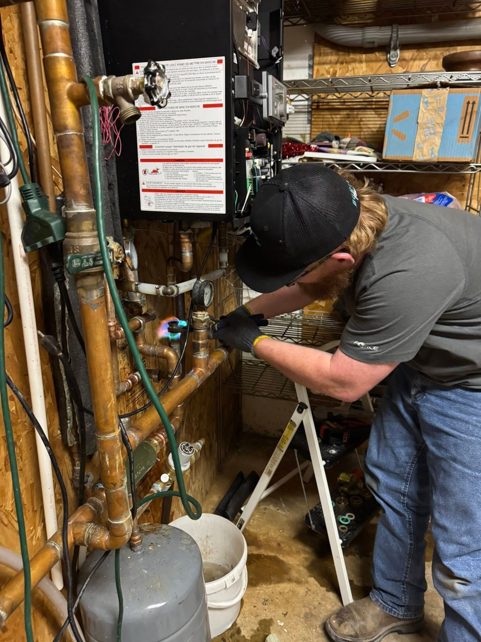 Plumber working on copper pipes in a basement; wearing hat, gloves, and jeans. Standing on a stepladder.