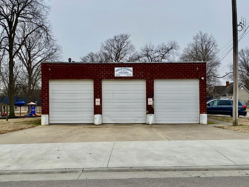 Red brick three-bay garage with white doors beside a street, with leafless trees in the background