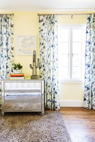 A bedroom with yellow walls , blue and white curtains , a mirrored nightstand , and a window.