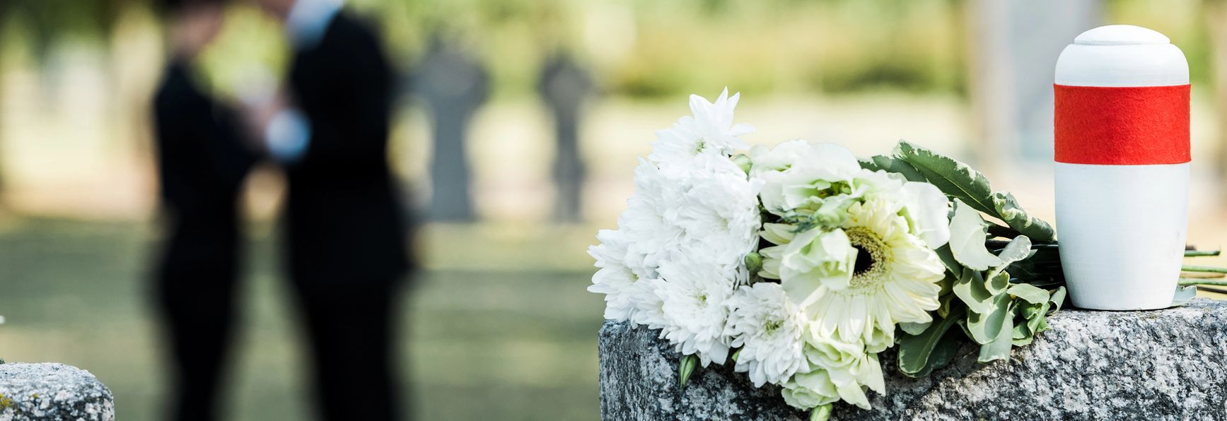 Scena di un cimitero con fiori e una lapide commemorativa; persone in lutto sullo sfondo.