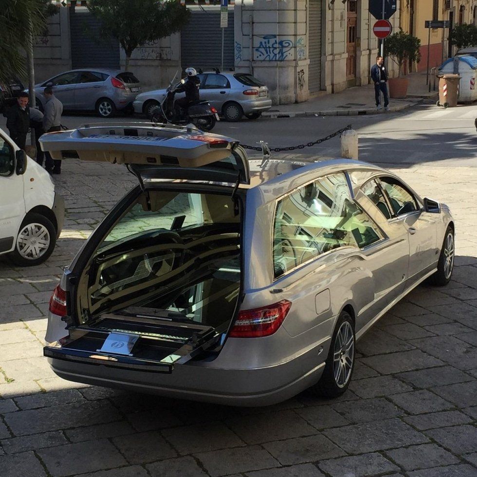 Carro funebre grigio con la portiera posteriore aperta, parcheggiato in una strada cittadina.