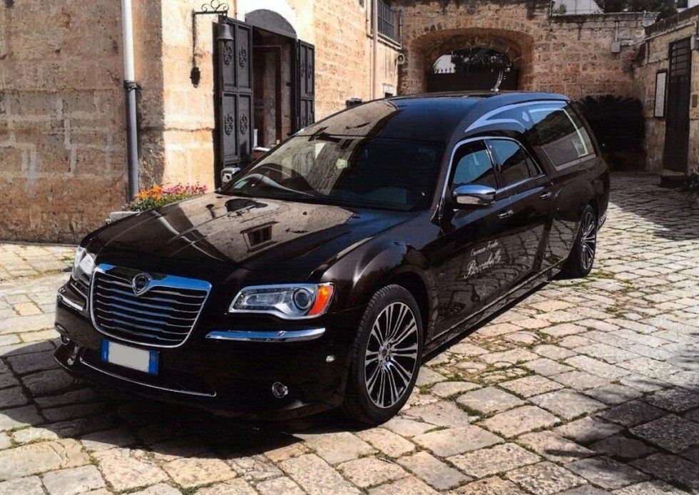 Carro funebre nero parcheggiato su una strada acciottolata, accanto a un edificio.