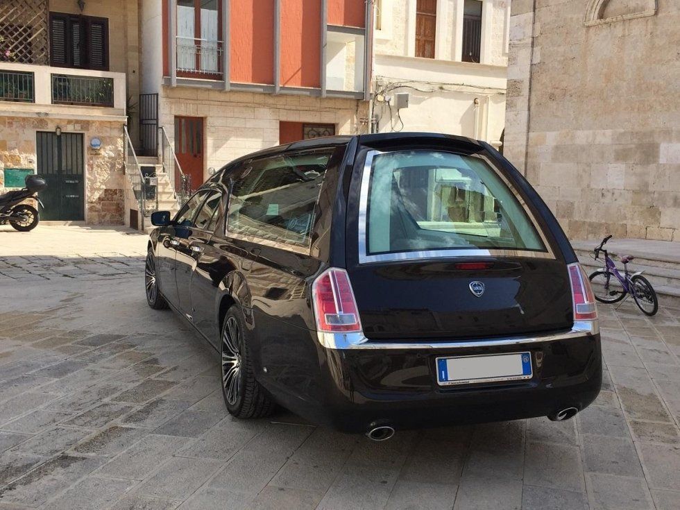 Carro funebre nero parcheggiato su una piazza acciottolata di fronte agli edifici.