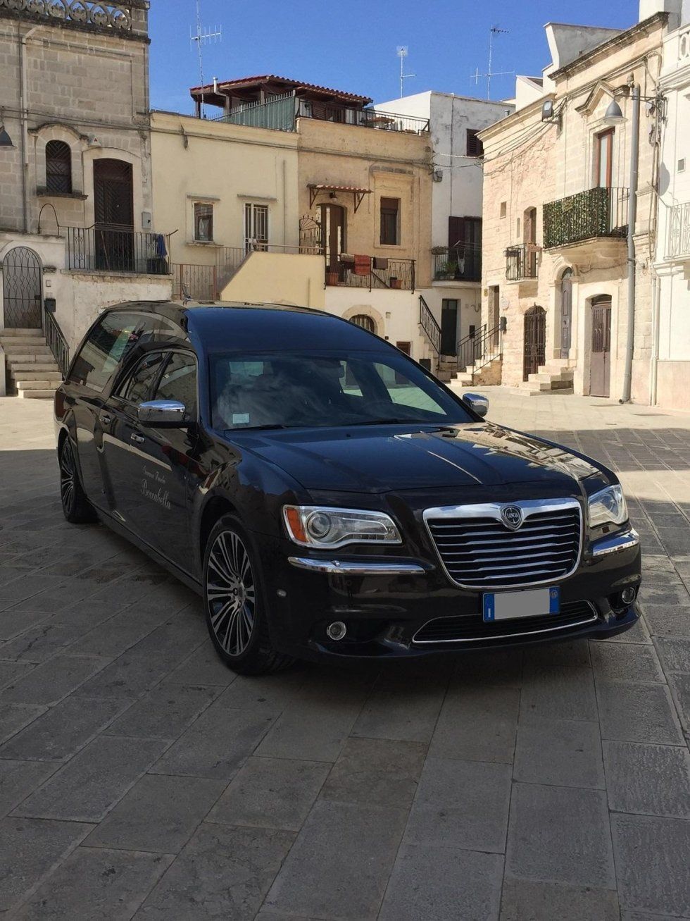Un carro funebre nero parcheggiato in una piazza acciottolata, circondato da edifici in pietra sotto un cielo soleggiato.