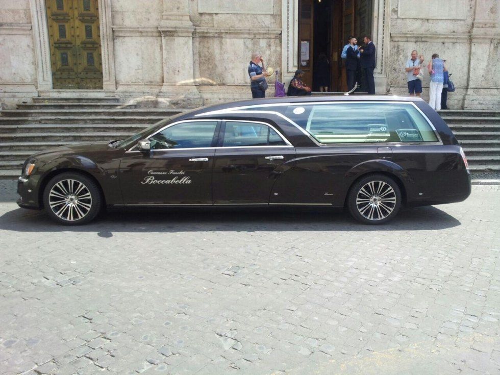 Carro funebre marrone scuro parcheggiato davanti a un edificio con gradini; persone in piedi nelle vicinanze.