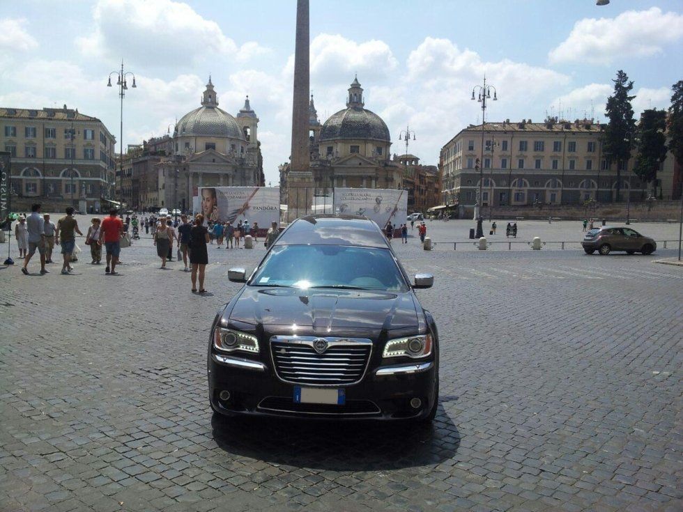 Auto scura parcheggiata in una piazza acciottolata; persone che camminano; edifici e obelisco sullo sfondo, giornata di sole.