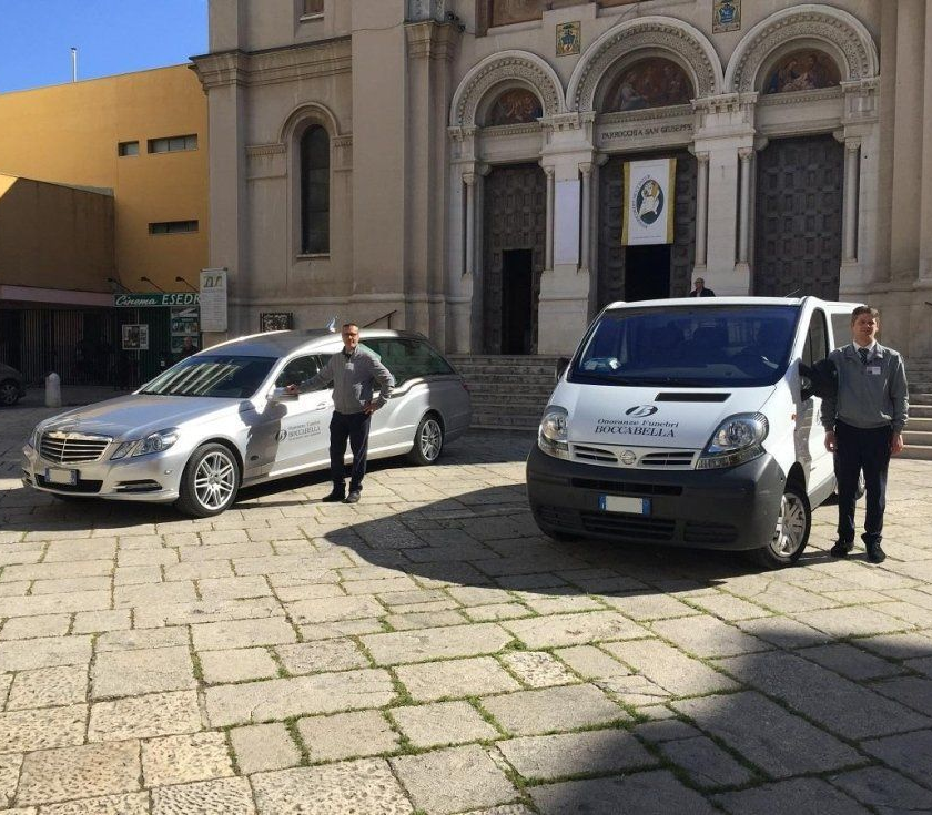 Due uomini sono in piedi accanto a un carro funebre e a un furgone di fronte a una chiesa.