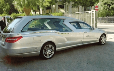 Carro funebre argentato parcheggiato, vista laterale, su una superficie asfaltata vicino a un edificio.