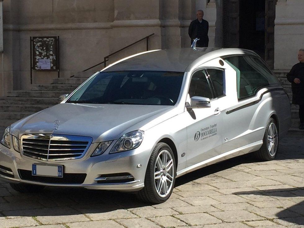 Carro funebre argentato parcheggiato davanti a un edificio, circondato da persone.