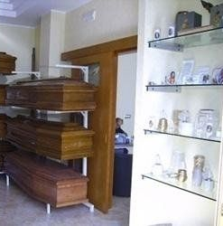 Interno di un'agenzia di pompe funebri con diverse bare esposte su scaffali. I ripiani in vetro ospitano urne e oggetti religiosi.