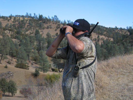 A man is looking through binoculars in a field with trees in the background