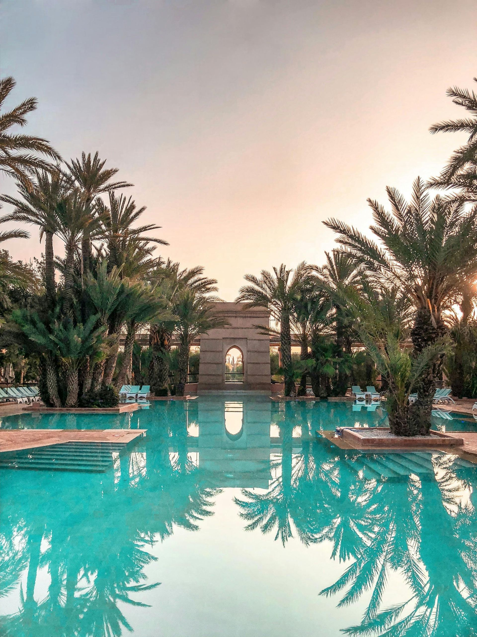 Swimming pool lined with palm trees reflecting in the turquoise water, with a stone archway in the background.