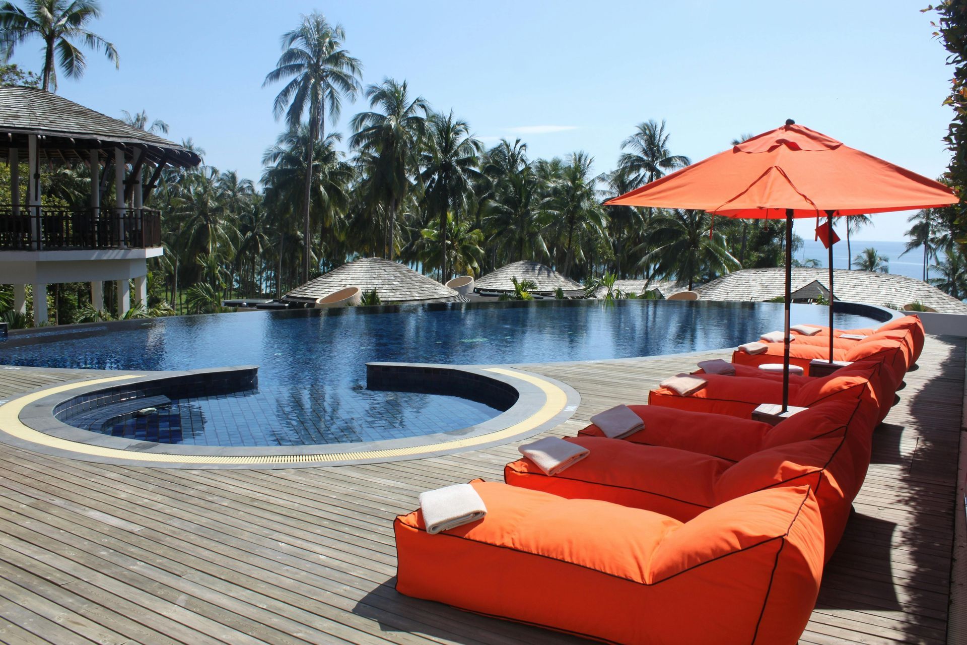 A pool with orange lounge chairs, umbrella, and clear blue water with palm trees in the background.