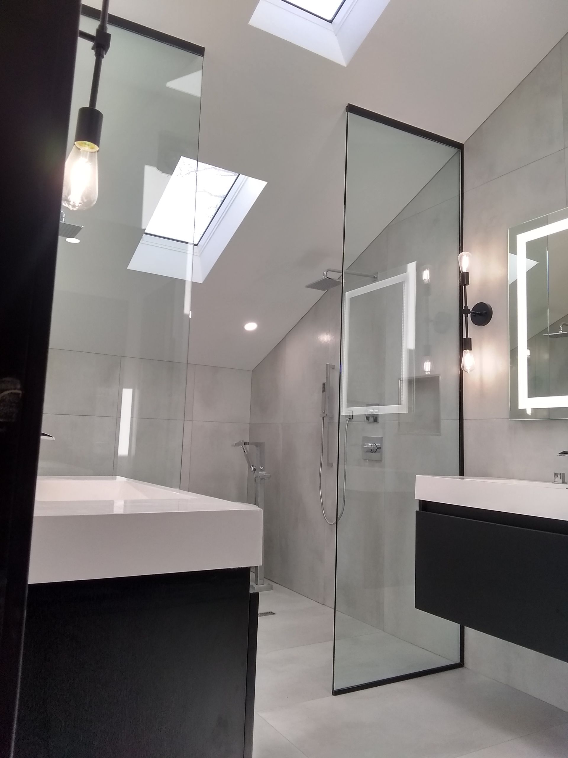 A bathroom with a walk in shower , sink , mirror and skylight.
