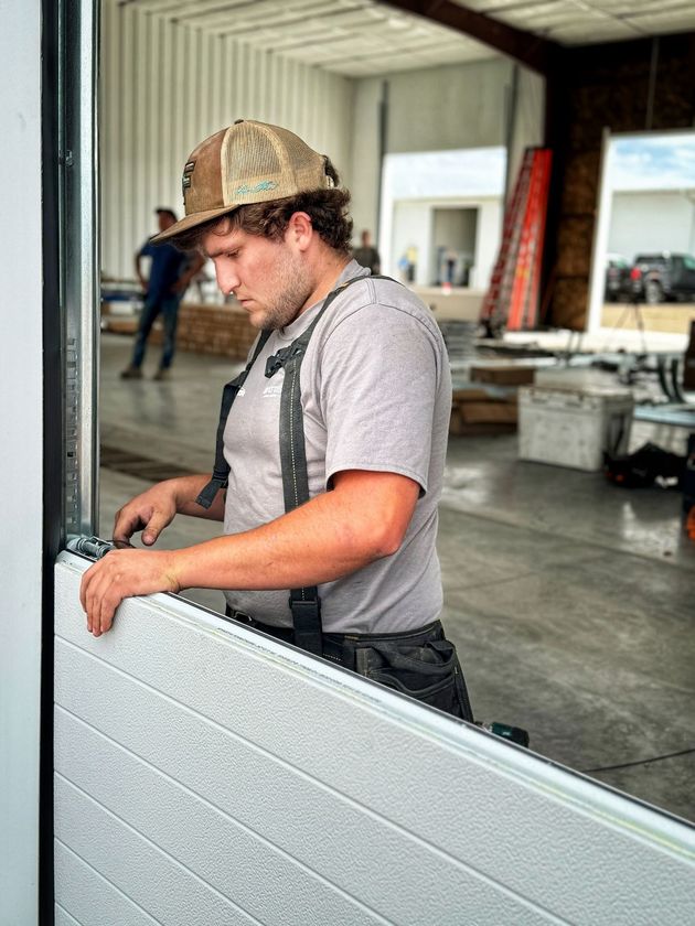A person wearing a tan cap and grey t-shirt installs a grey panel on a large industrial door inside a workshop.