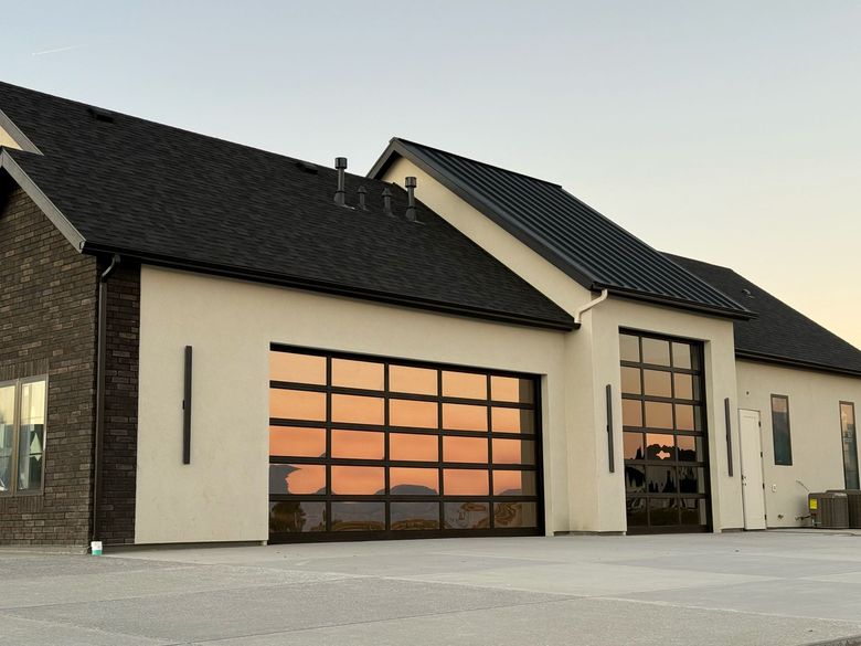 A modern house with cream stucco walls, dark roof tiles, and two large, glass-paneled garage doors reflecting the sunset.