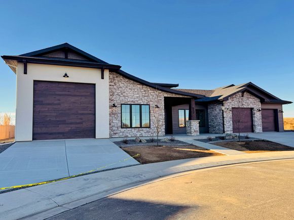 A single-story suburban house with stone and stucco exterior, dark garage doors, and a paved driveway under a blue sky.