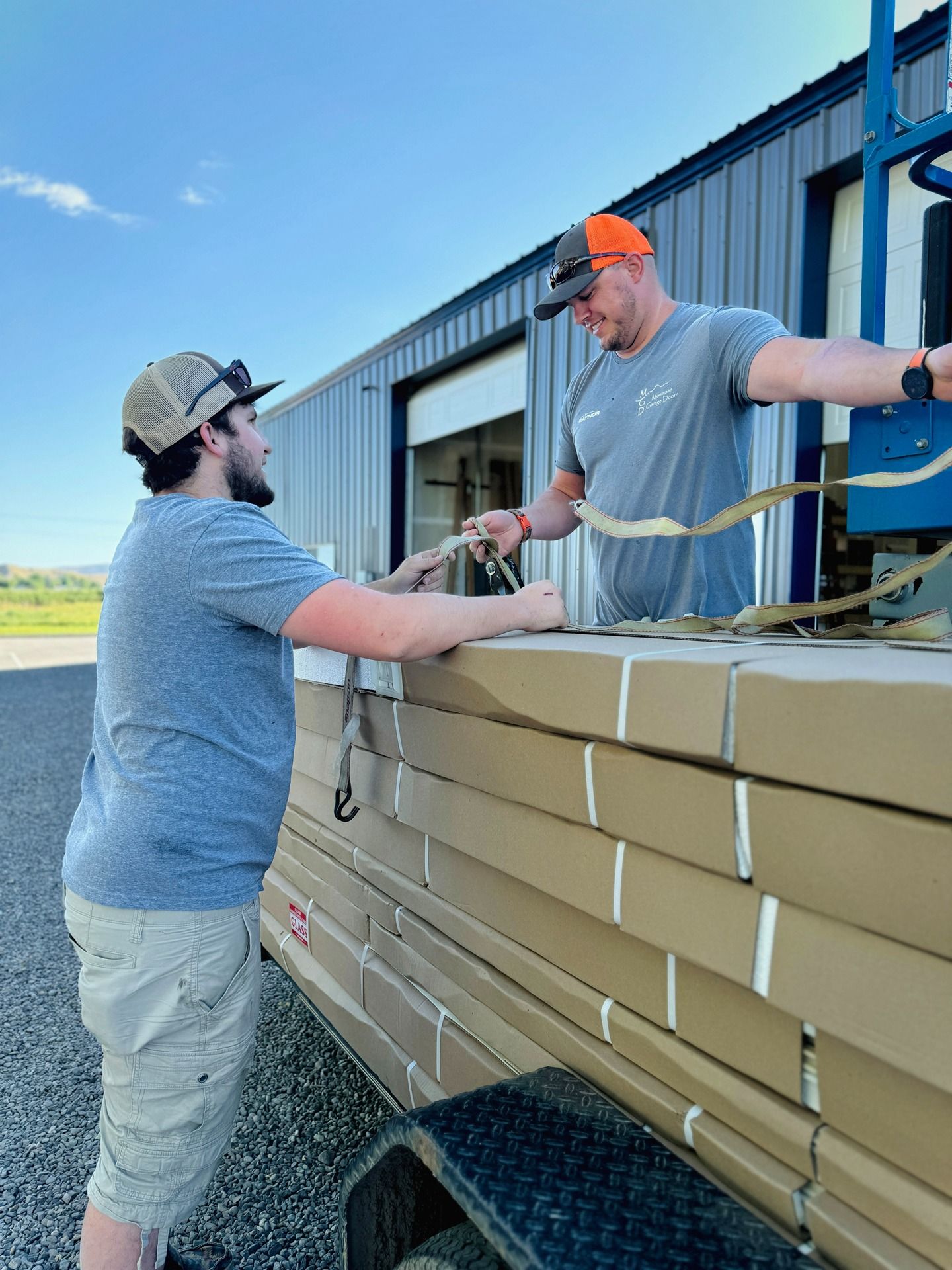 Two people wearing hats secure a large stack of flat, tan-colored packages onto a flatbed trailer outdoors.