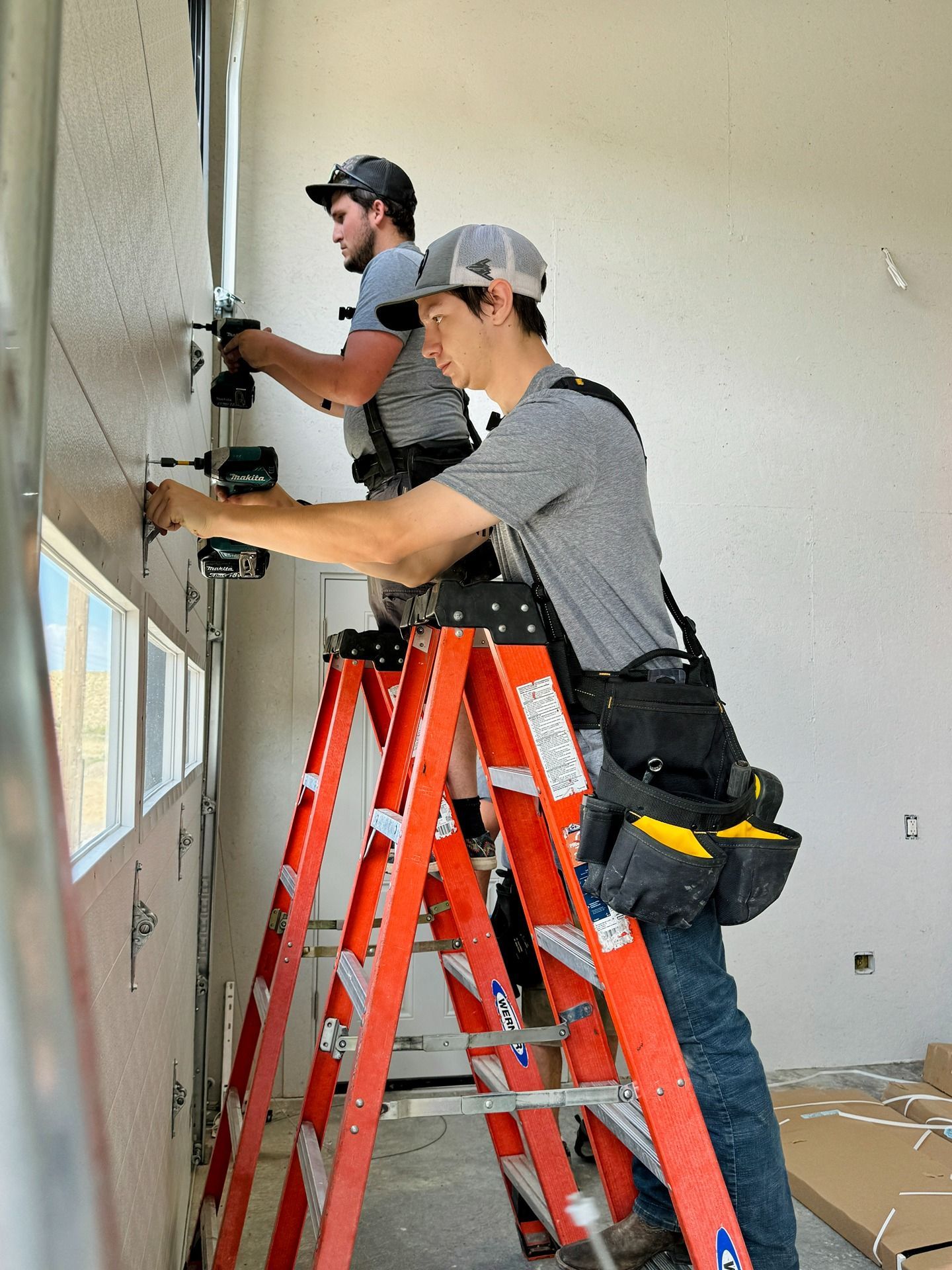 Two technicians in gray shirts and hats stand on red A-frame ladders, using power drills to install a garage door panel.