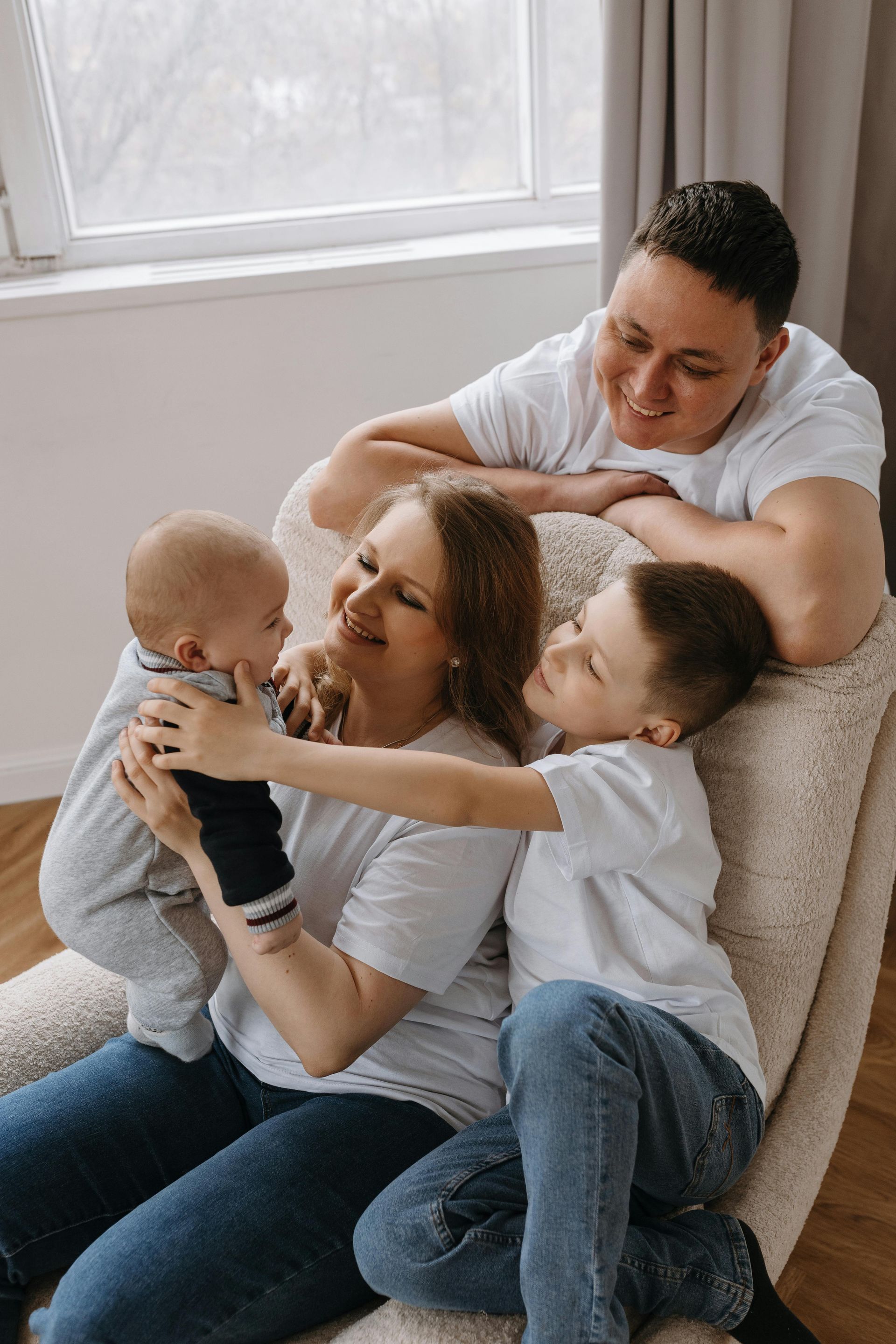 Family hugging on a couch by a window, smiling and embracing two small children