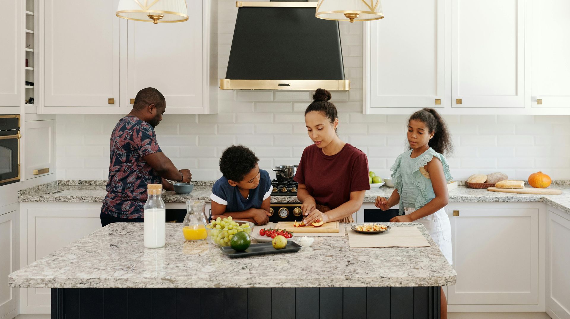 Family preparing food together in a bright white kitchen around a large island with fruits and vegetables.
