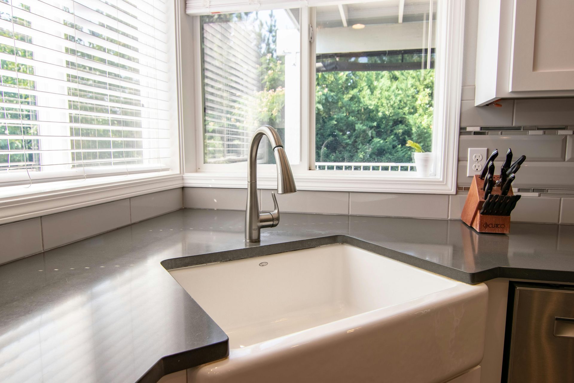 Bright kitchen sink with gray countertop, white cabinets, and large windows overlooking greenery.