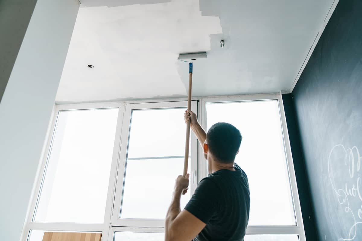 A person painting a white ceiling with a long-handled roller in a room with large windows.