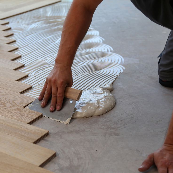 A person uses a notched trowel to spread adhesive onto a concrete floor before laying wood planks.