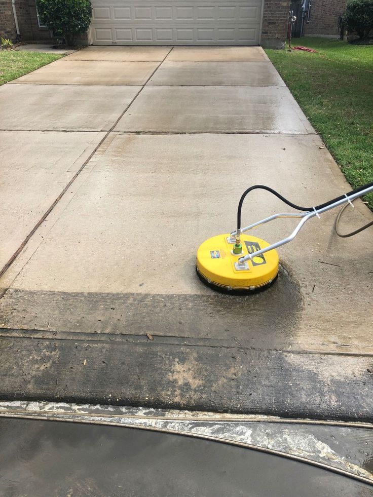 A yellow pressure washer surface cleaner cleans a concrete driveway, showing a clear contrast between dirty and clean areas.