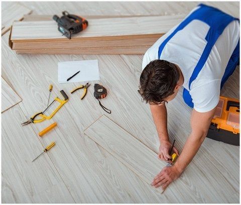 A person in blue coveralls kneels on a light wood floor, using a pencil and tape measure to mark a plank for installation.