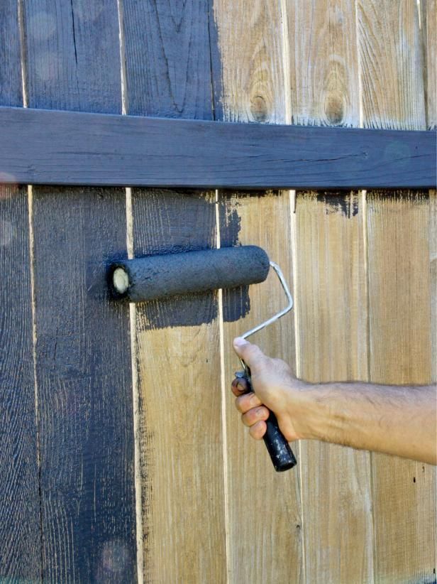 A hand uses a paint roller to apply dark stain to vertical wooden fence boards.