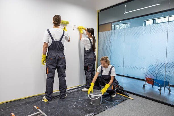 Three workers in dark gray jumpsuits and yellow gloves paint a white wall inside a modern office with glass partitions.