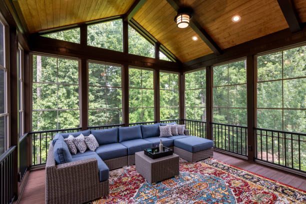 A screened-in porch with a blue sectional sofa, a square coffee table, and a patterned rug overlooking a forest.
